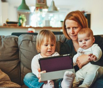Eine Frau sitzt mit einem Baby und einem Kleinkind auf dem Sofa. Sie schauen sich etwas auf einem Tablet an. Zum Thema: Kinder und Fernsehen
