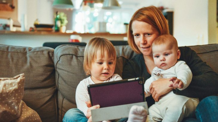 Eine Frau sitzt mit einem Baby und einem Kleinkind auf dem Sofa. Sie schauen sich etwas auf einem Tablet an. Zum Thema: Kinder und Fernsehen